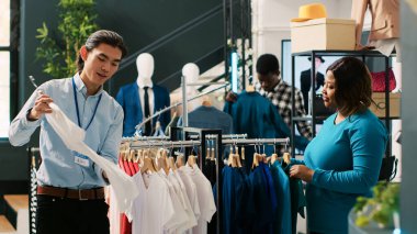 Mağaza müdürü gündelik kıyafetlerle müşterilere yardım ediyor, Afro-Amerikan kadın moda ürünler alıyor. Asyalı bir çalışan modern butikte şık kıyafetler ayarlıyor. Moda konsepti