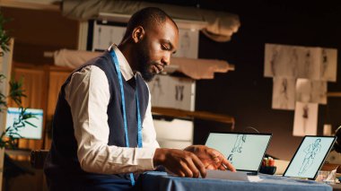 Tabletteki tasarımları inceleyen terzi atölyedeki kıyafetlerin çizimlerini karşılaştırıyor. Moda tasarımcısı terzilik aletleri ve aletleri kullanarak giyim markası üretiyor. El kamerasıyla..