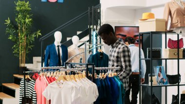 Afro-amerikan alışverişçi, temel gömleği kontrol ediyor, giyim mağazasındaki kumaş ürünlerini analiz ediyor. Modern butikte alışveriş manyağı bir müşteri şık kıyafetler alıyor. Moda konsepti