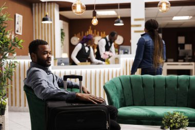 Genç Afrikalı Amerikalı erkek yolcu otel girişini bekliyor, yanındaki bavulla koltukta oturuyor, rahat dinlenme odasında dinleniyor. Lobide bagajı olan zenci bir işadamı.