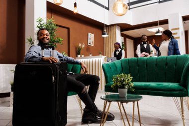 Mutlu gülümseyen Afrikalı Amerikalı adam otel lobisinde bavuluyla oda bekliyor. Pozitif milenyum siyah erkek turist kontrol saatini beklerken koltukta dinleniyor.