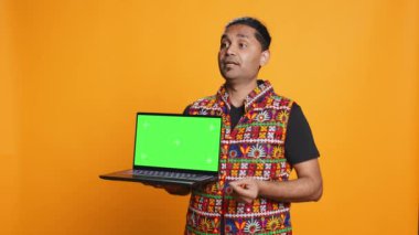 Teknoloji içeriği yaratıcısı yeşil ekran laptop kullanarak pazarlama yapıyor, stüdyo arka planında izole edilmiş. Hintli adam elinde boş fotokopi defteri tutuyor, inceleme yapıyor, kamera B
