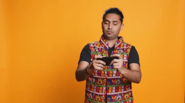 Geleneksel giyimli neşeli Hintli insan akıllı telefonuyla video oyunlarıyla eğleniyor. Neşeli oyuncu telefondan keyif alıyor, çevrimiçi eğleniyor, stüdyo arkaplanı, kamera B