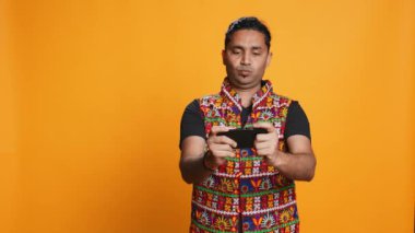 Akıllı telefondaki video oyunlarıyla eğlenen mutlu Hintli adam boş vakitlerin tadını çıkarıyor. Oyuncu telefonda oyundan zevk alıyor, düşmanları yenerken eğleniyor, stüdyo arka planında izole edilmiş, kamera B