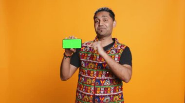 Gülümseyen Hintli adam cep telefonuyla yeşil ekran ekranı sunuyor, stüdyo arka planında izole edilmiş. Geleneksel giysiler içinde, boş fotokopi odası modeliyle promosyon yaratan kişi, kamera B