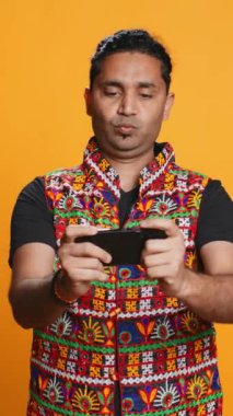 Dikey videolu mutlu Hintli adam akıllı telefondaki video oyunlarıyla eğleniyor, boş zamanların tadını çıkarıyor. Oyuncu telefonda oyundan zevk alıyor, düşmanları yenerken eğleniyor, stüdyo arka planında izole edilmiş, kamera B