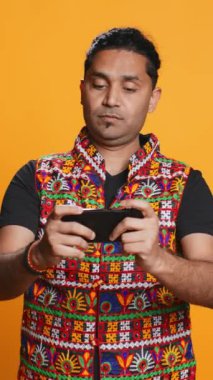 Geleneksel giysiler içinde video oyunlarıyla akıllı telefondan eğlenen dikey video çeken neşeli Hintli insan. Neşeli oyuncu telefondan keyif alıyor, çevrimiçi eğleniyor, stüdyo arkaplanı, kamera B