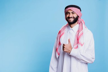 Geleneksel kıyafetler giymiş Müslüman bir adam, neşeli bir yüz ifadesiyle başparmağını gösterirken. Gülümseyen İslami kişi onaylıyor ve kameraya bakıyor.