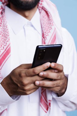 Arap adam cep telefonunu elinde tutuyor, sosyal medya ağını kaydırıyor ve çevrimiçi yakın çekim mesajı atıyor. Geleneksel kıyafetler içindeki Müslüman kişi akıllı telefon ve internet sörfü kullanıyor