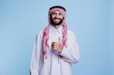 Arap adam geleneksel İslami kıyafetler giyiyor. Baş parmağıyla poz veriyor. Onaylama ve uzlaşma portresi. Thombe ve Gutra 'da gülümseyen müslüman bir jest gibi görünüyor.