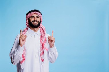 Heyecanlı müslüman satış elemanı stüdyo portresi için poz verirken yukarı doğru bakıyor. Geleneksel kıyafetler giymiş neşeli gülümseyen Arap adam ortaya çıkıp kameraya bakıyor.