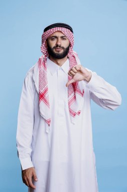 Beyaz mantolu ve başörtülü Müslüman bir adam başparmağıyla poz veriyor ve kendinden emin bir ifadeyle kameraya bakıyor. Ciddi Arap portresi, fikir ayrılığı anlamına geliyor.