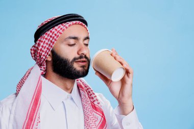 Müslüman adam geleneksel İslami kıyafetler giyip kağıt bardaktan kahve içiyor. Arap bir adam tobe ve başörtüsü takmış elinde kupa tutuyor ve sıcak yemek servisinin tadını çıkarıyor.