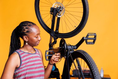 Sports-loving woman expertly adjusting broken bicycle crank arm with specialized multitool. Young african american lady carefully examining and fixing bike components with professional allen key. clipart