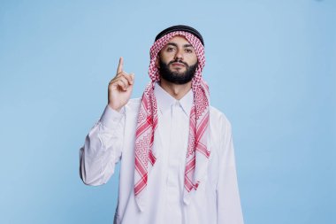 Müslüman adam Arap kıyafetleri giyip, parmak stüdyosunun portresini göstererek ürün reklamı yapıyor. Geleneksel bornoz giymiş İslami kişi yukarı doğru bakıyor ve kameraya bakıyor.