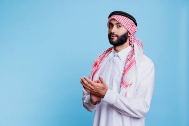 Geleneksel kıyafetler giymiş ciddi bir Arap adam alkışlıyor ve destek portresi gösteriyor. Müslüman kişi el çırpıyor ve kendine güvenen yüz ifadesiyle kameraya bakıyor.