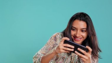 Hintli bir kadın cep telefonuyla oyun oynuyor manzara modunda, boş zamanların tadını çıkarıyor. Oyuncu cep telefonundan zevk alıyor, eğleniyor, stüdyo arka planında izole edilmiş, kamera B