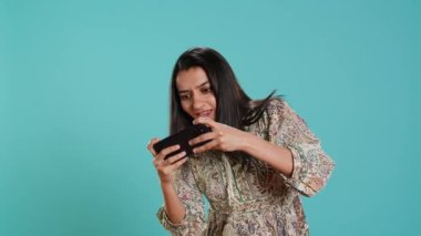 Akıllı telefonda video oyunları oynayan mutlu bir kadın düşmanları yendikten sonra kutlama yapıyor, el hareketi yapıyor. Oyuncu telefon ekranı araklıyor. Maçı kazanmak için heyecanlanıyor. Stüdyo arka planı, kamera A.