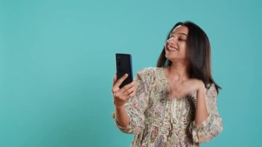 Mutlu kadın, arkadaşlarıyla sohbet etmek için internetten akıllı telefondan arkadaşça jestler yapıyor. Telekonferansta gülümseyen Hintli insan cep telefonu, stüdyo arka planı, kamera A 'yı kullanıyor.