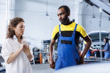 BIPOC tamircisi araba tamirhanesinde müşteriye yardım ediyor. Garaj çalışanı kadınla otomobil parçalarına bakıyor, teftiş sırasında aracının motorunu tamir ediyor.