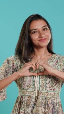 Neşeli, dost canlısı Hintli kadının elleriyle kalp sembolü hareketi yaptığı dikey video görüntüsü. Sevgi dolu davranışlar sergileyen neşeli bir insan, stüdyo arka planında izole edilmiş, kamera A