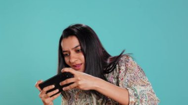 Akıllı telefonda video oyunları oynayan mutlu bir kadın düşmanları yendikten sonra kutlama yapıyor, el hareketi yapıyor. Oyuncu telefon ekranını araklıyor. Maçı kazanmaktan heyecan duyuyor. Stüdyo arka planı, kamera B