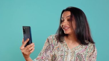 Mutlu kadın, arkadaşlarıyla sohbet etmek için internetten akıllı telefondan arkadaşça jestler yapıyor. Telekonferansta gülümseyen Hintli insan cep telefonu, stüdyo arka planı, kamera B kullanarak konuşuyor.