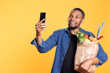 Taze ürünlerle dolu bir çantayla fotoğraf çeken gülümseyen pozitif adam etik kaynaklı mallardan memnun. İyimser arkadaş cep telefonu uygulamasından fotoğraf çekiyor..