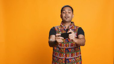 Akıllı telefondaki video oyunlarıyla eğlenen mutlu Hintli adam boş vakitlerin tadını çıkarıyor. Oyuncu telefonda oyundan zevk alıyor, düşmanları yenerken eğleniyor, stüdyo arka planında izole edilmiş, kamera B
