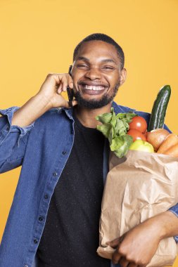 Afrika kökenli Amerikalı karizmatik yetişkin sohbetleri stüdyodaki mobil cihazında, yerel süpermarketten gelen poşetlerle dolu bir poşet taşıyor. Neşeli arkadaş alışveriş sırasında telefona cevap verir..