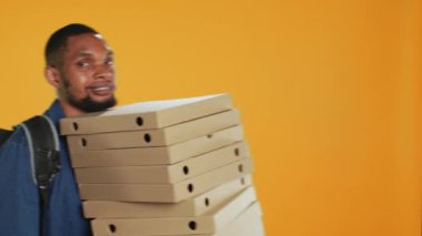 Afrikalı Amerikalı pizzacı, stüdyodaki müşterilere yemek siparişi vermek için elinde bir yığın pizza kutusu tutuyor. Dost canlısı servis elemanı paket servis paketi taşıyor. Kamera A.