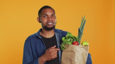 Afro-amerikalı bir insan, bir kese kağıdında depolanmış sebze ve meyveleri işaret ederek, sağlıklı beslenme ve vejetaryen beslenmesi için çevre dostu tavsiyelerde bulunuyor. Kamera A.