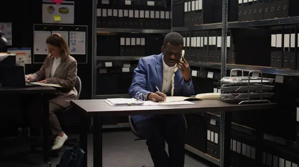 stock image African american police officer conducts private investigation, evaluates evidence, and places important phone calls. Male law enforcement agent uses landline to gather classified witness information.