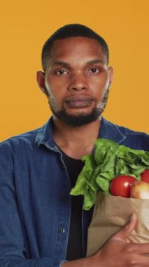 Vejetaryen birinin dikey video portresi. Taze sebzelerle dolu bir poşet taşıyor. Yerel çiftçiliği ve ev yapımı ürünleri destekliyor. Erkek model biyoloji süpermarketinde alışveriş çılgınlığının tadını çıkarıyor. Kamera.