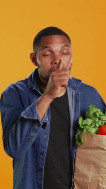 Dikey Video Afrikalı Amerikalı gizemli adam sessiz sessiz sessiz hareketler yapıyor, parmakları dudaklarında, yerel organik ürünler hakkında bir sır saklıyor. Vejetaryenlik konusunda özel olan kişi