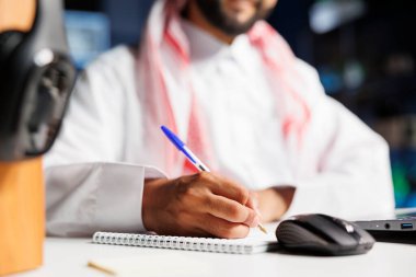 Masada oturan Orta Doğulu bir adam tarafından kullanılan defter ve kalemin yakın plan fotoğrafı. Araştırma notları yazan Müslüman adama odaklan..