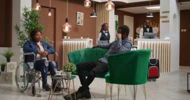Tekerlekli sandalyede oturup maluliyet dostu otel lobisine girerken Afrikalı Amerikalı bir misafir adamla konuşuyor. Yurt dışına seyahat eden bir çift, seyyar turizm sektöründe azalma.