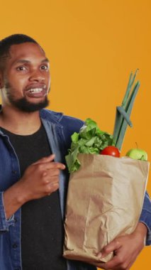 Dikey Videolu Afrikalı Amerikalı insan, bir kese kağıdında depolanmış meyve ve sebzeleri işaret ederek sağlıklı beslenme ve vejetaryen beslenmesi için çevre dostu tavsiyelerde bulunuyor. Kamera B.