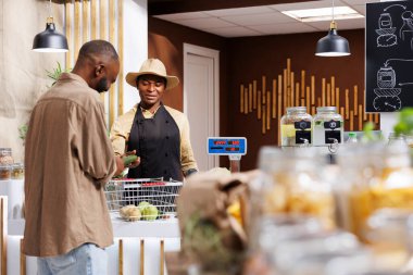 Çevreye duyarlı Afrikalı Amerikalı müşteriler yerel bir süpermarkette organik, yerel kaynaklı meyve ve sebzeler hakkında sorular soruyorlar. Kasada müşterisine yardım eden şapkalı bir satıcı..