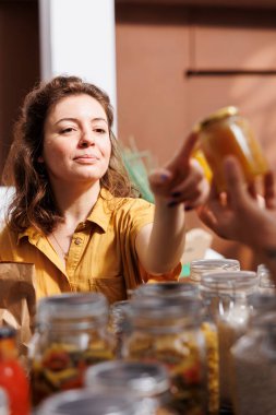 Storekeeper showing woman shopping in zero waste grocery store organic nutritious honey in reusable packaging. Vendor in ethically conscious shop recommending food item to customer clipart