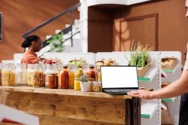 Beyaz ekranlı dizüstü bilgisayar, cam şişeler ve kavanozlarla dolu ahşap masanın üzerine yerleştiriliyor. Arka planda, satıcı izole edilmiş kromatonlu mini bilgisayar kullanırken bir kadın dükkana bakıyor..