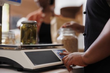 Önlüklü Afro-Amerikalı bir birey, bakkal kasasında müşteriye yardım ediyor. Çevre dostu biyo-gıda pazarındaki kadın satıcı, cam turşu kavanozunu dijital ölçekte tartıyor.