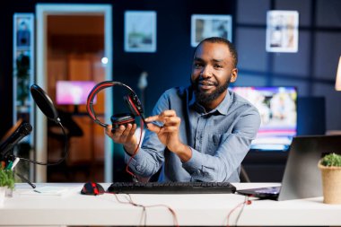 İnternet kanalı için loş ışıklı ev stüdyosunda video kaydeden Afrikalı Amerikalı bir adam abonelere oyun kulaklığı sunuyor. İzleyicilere ses dinleme aygıtı gösteren ortam yıldızı