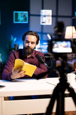 Stüdyoda deneyimli ses sanatçısı sesli kayıt üzerinde çalışıyor, kitaptan okuyor, profesyonel mikrofonla ses kaydediyor ve yüksek kaliteli ekipman kamerasıyla çekim yapıyor.