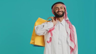 Stüdyoda bir sürü alışveriş çantası taşıyan Müslüman bir adam, kıyafet ve mal satın alma konusunda başarılı olduktan sonra özgüvenle poz veriyor. Arap kişi perakende mağazasında indirimli alışveriş merkezinden ürün satın alıyor.