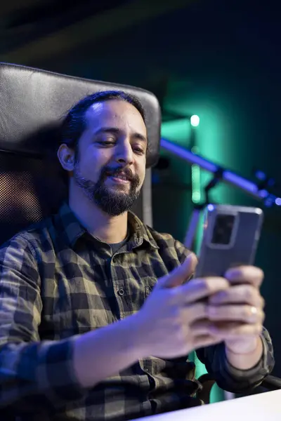 stock image Cheerful male student is utilizing his mobile device to browse social media while seated at his desk. Bearded man with a smile answers texts while using his smartphone for surfing the internet.