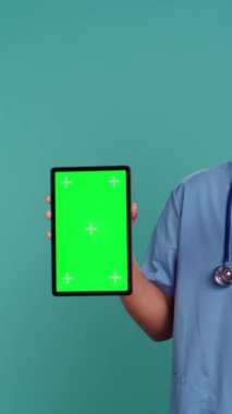 Erkek hemşirenin dikey video portresi yeşil ekran tabletinde tıbbi talimat videosunu gösteriyor. Hastane çalışanı, elinde kroma anahtarı var. Mavi stüdyo arka planında izole edilmiş. Kamera B.