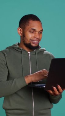 Dikey videolu gülümseyen adam dizüstü bilgisayarda kayıyor, ürünleri ödemek için internet ve kredi kartı kullanıyor. Afrikalı Amerikalı mutlu bir şekilde online alışveriş sitelerine giriyor, stüdyo arka planında izole edilmiş.