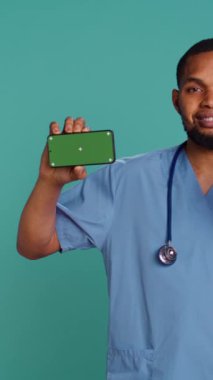 Neşeli erkek hemşirenin dikey video görüntüsü izole ekran akıllı telefonuna işaret ediyor. Radyasyon uzmanının elinde kroma anahtar telefon, mavi stüdyo arka plan, kamera B