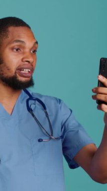 Video konferansında cep telefonu kullanan, insanları selamlayan dikey video hemşire. Stüdyoda izole edilmiş, akıllı telefon kullanan hastalarla çevrimiçi teletıp görüşmesi yapan sağlık uzmanı.